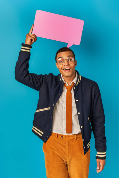 smiling african american student in trendy jacket and eyeglasses holding speech bubble on blue - Foto, afbeelding