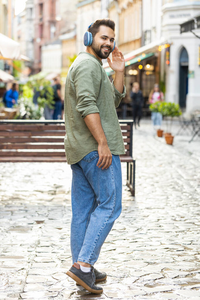 Rear view of man tourist in wireless headphones walking through the street outdoors, listening energetic music, dancing turning to camera and smiling. Back view of guy traveler in urban city. Vertical - 写真・画像