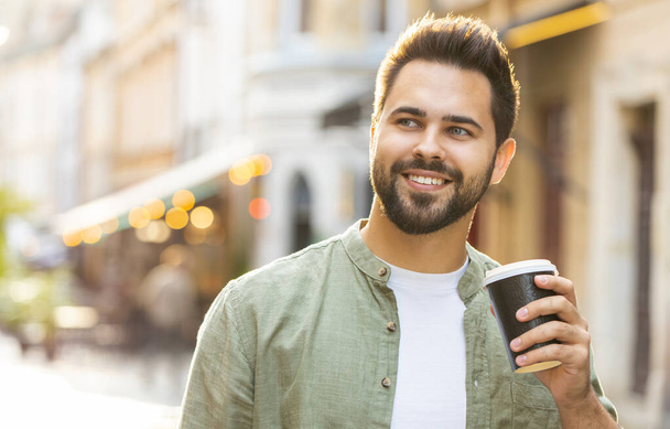 Счастливый бородатый молодой человек наслаждается утренним кофе горячим напитком и улыбается на открытом воздухе. Расслабляюсь, делаю перерыв. Парень, гуляющий по городской солнечной улице, пьющий кофе на вынос. Городской образ жизни снаружи - Фото, изображение