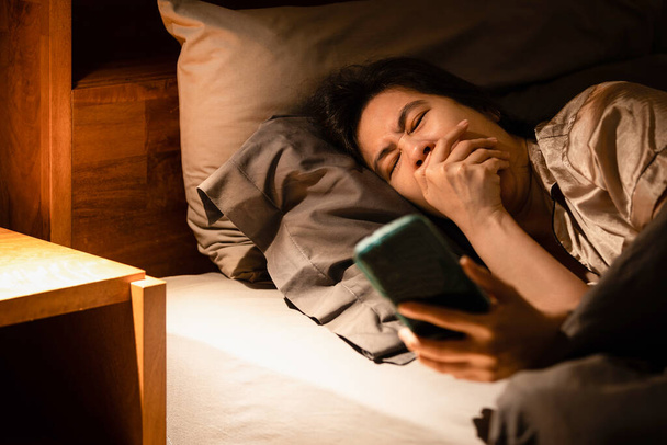 Schläfrige Frau, die im Bett liegt, abhängig von ihrem Handy, ständig gähnt, sich müde fühlt, ihre schweren Augenlider sich schwer tun, offen zu bleiben, ihren schläfrigen Zustand bekämpft, Symptome von Schlafstörungen oder Racheschlaf - Foto, Bild
