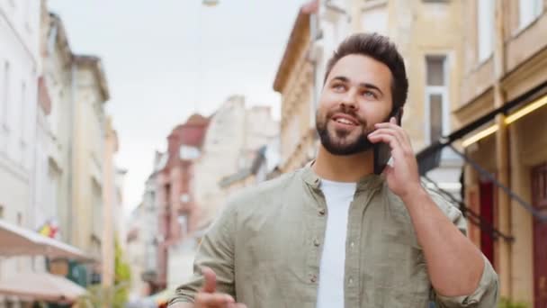 Onnellinen nuori mies turisti ottaa etäkeskustelun kommunikoida puhuminen älypuhelimella. Parrakas kaveri puhuu puhelimessa odottamattomista hyvistä uutisista kävelemässä kaupunkikadulla. Kaupunkien elämäntavat ulkona - Materiaali, video