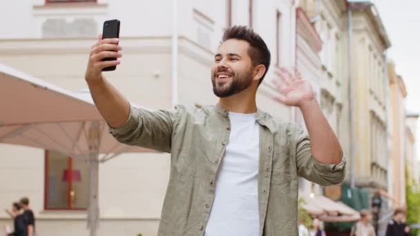 Bearded giovane blogger scattare selfie su smartphone, comunicare videochiamata online con gli abbonati o amici di famiglia, registrare storie per i social media vlog all'aperto. ragazzo a piedi in città strada - Filmati, video