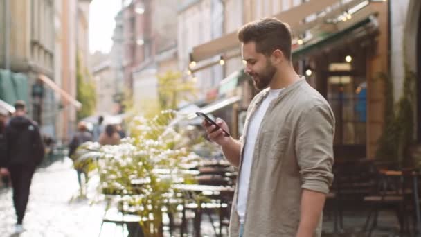 観光客の旅行者は、屋外のモバイルナビゲータアプリで地図上の方法を検索するためのスマートフォンの入力テキストメッセージを使用して若い男にひげを生やした。男歩くパスによって都市太陽の街の背景 - 映像、動画