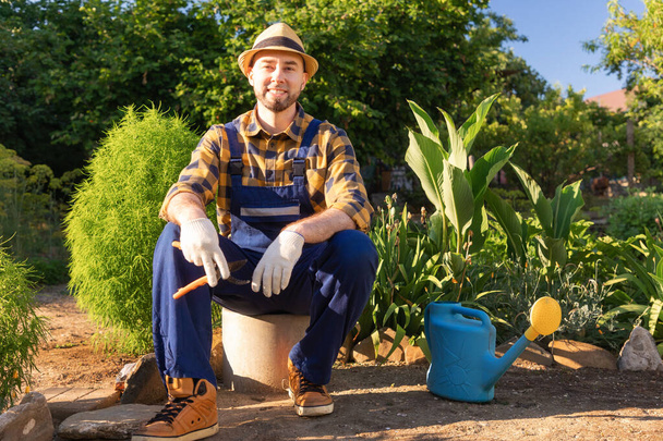 Happy bearded, handsome man in overalls and a straw hat sits and relax, holding a pruner. In the background is a backyard and a kitchen-garden. The concept of home gardening. - Фото, изображение