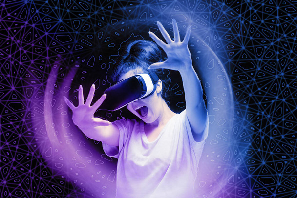 Zszokowana młoda kobieta w okularach VR obawia się 3D symulacji rzeczywistości wirtualnej. Ciemne tło z neonową siatką i hologramem. Koncepcja metawszechświata i cyberprzestrzeni. - Zdjęcie, obraz