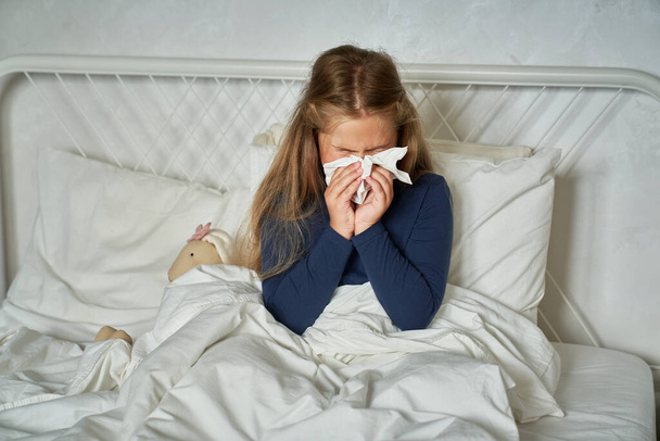 Krankes Mädchen im Grundschulalter pustet Nase, während es auf dem Bett sitzt - Foto, Bild