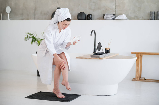 Woman in bathrobe and towel applying body lotion or balm on legs. Skincare after shaving, epilation, depilation or hair removal. Medical gel for treatment of varicose veins disease or edema. - Photo, Image