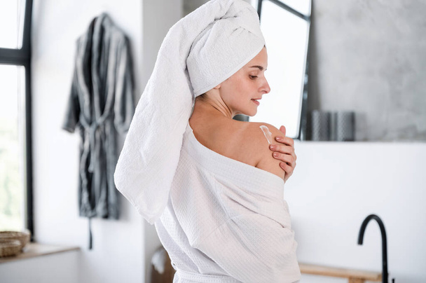 Back view of woman in towel and robe applying moisturizer cream on shoulder skin after shower. Female performing daily skincare routine in bathroom at home. Body care concept - Photo, Image