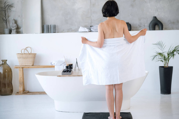 Vista posterior de la mujer envuelta en toalla blanca esponjosa de pie cerca del baño en un estilo minimalista brillante. Preparación para los procedimientos de spa en casa, lavado. Pasar tiempo en el baño. - Foto, Imagen