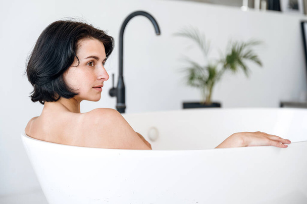 Portret van een jonge vrouw die wegkijkt terwijl ze een heet bad neemt in de badkamer. Wellness, schoonheid en lichaamsverzorging concept. Ontspanning en rust thuis. Kopieerruimte. - Foto, afbeelding