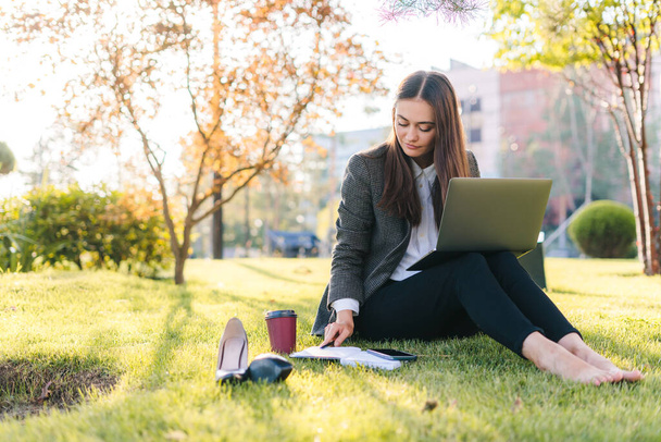 Mujer joven y despreocupada freelancer que usa ropa casual usando un portátil mientras está sentado en la hierba con una taza de café desechable. Adolescente estudiante chica de navegación - Foto, imagen