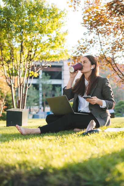 Jeune femme aux cheveux bruns buvant du café à emporter et utilisant un ordinateur portable assis sur de l'herbe verte. Utilisation de l'ordinateur. Enseignement à distance en ligne et boutiques - Photo, image