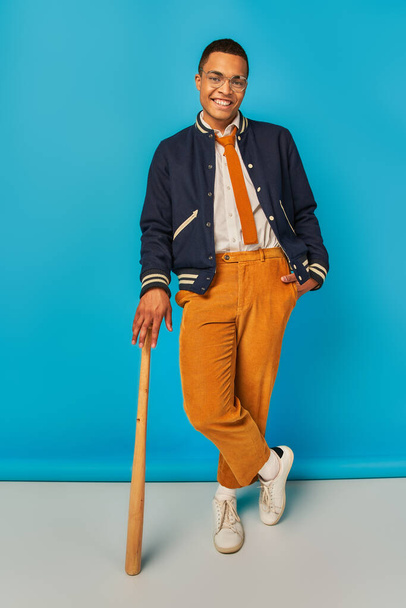 usmívající se africký americký student v bundě a oranžové kalhoty pózování s baseballovou pálkou na modré - Fotografie, Obrázek