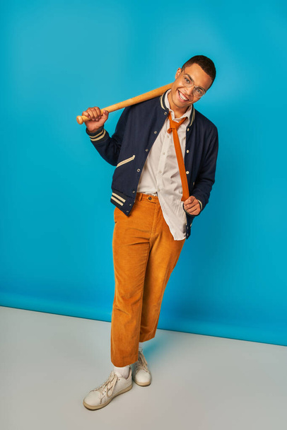 cheerful and stylish african american student with baseball bat pulling tie on blue - Photo, image