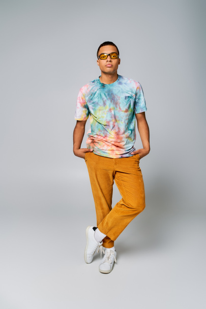 trendy african american man with hands in pockets on grey, orange pants, sunglasses, tie-dye t-shirt - Photo, Image