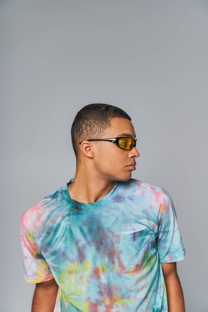youthful african american man in sunglasses and tie-dye t-shirt looking away on grey - Foto, Imagem
