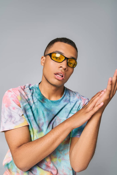 youthful, stylish african american man in tie-dye t-shirt and sunglasses looking at camera on grey - Fotoğraf, Görsel