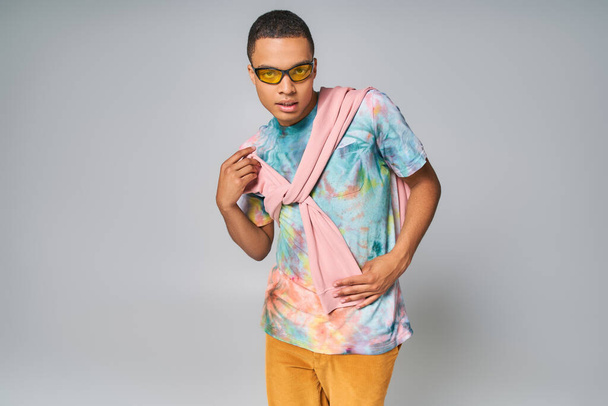 african american man in sunglasses and tie-dye t-shirt posing with hand on hip on grey - Fotografie, Obrázek
