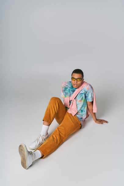 trendy african american man sitting on grey, sunglasses, orange pants, tie-dye t-shirt - Фото, зображення