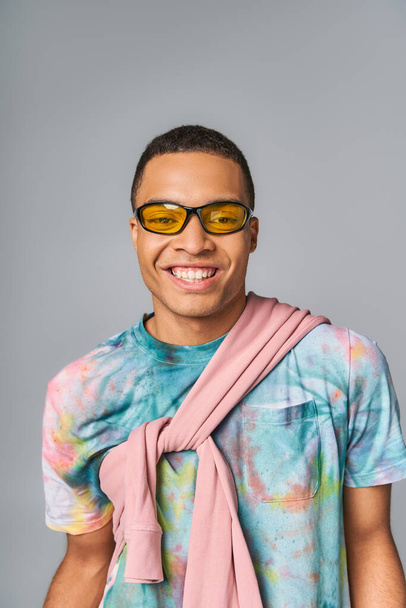 happy african american man in sunglasses and tie-dye t-shirt looking at camera on grey - Фото, изображение