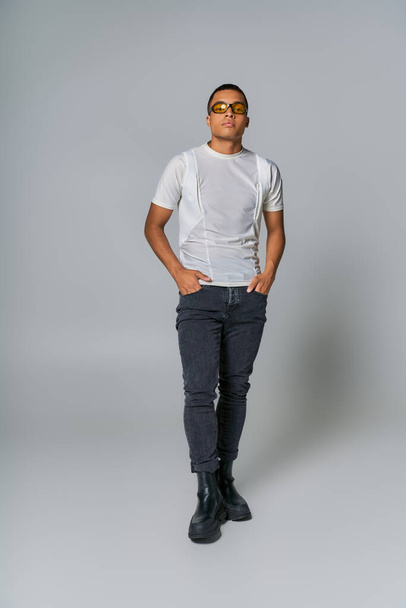 trendy african american man in sunglasses, t-shirt, jeans looking at camera on grey - Fotó, kép