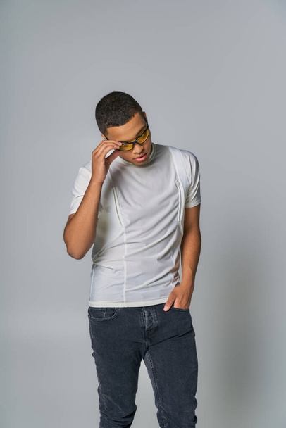 hombre afroamericano de moda en camiseta y jeans, con la mano en el bolsillo, ajustando las gafas de sol en gris - Foto, Imagen