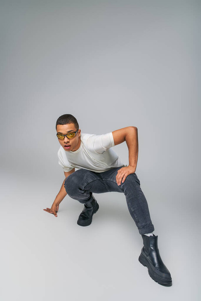 modern style african american guy in t-shirt, jeans and sunglasses looking at camera on grey - Foto, Imagem