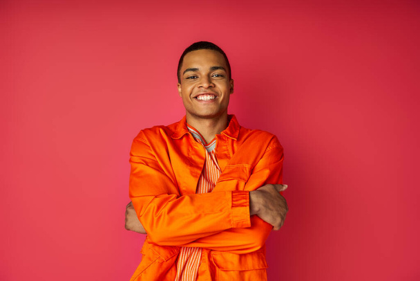 happy african american man in orange shirt, with folded hands, looking at camera on red - Foto, Imagen
