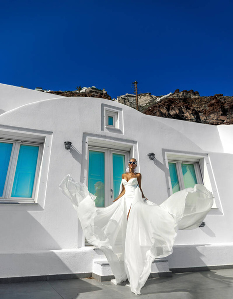 Stylish elegant woman in white long fluttering wedding dress posing on Santorini landscape with white buildings and beautiful pink flowers - Photo, Image