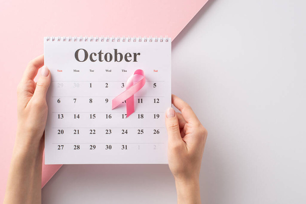 Sostenere la consapevolezza del cancro al seno in ottobre. Vista dall'alto delle mani che tengono un calendario e un nastro rosa su sfondo bianco e rosa pastello separato, fornendo spazio su copyspace per scopi testuali o pubblicitari - Foto, immagini