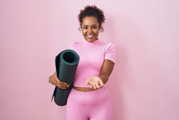 Junge hispanische Frau mit lockigem Haar hält Yogamatte über rosa Hintergrund lächelnd fröhlich bietet Handfläche Hand gibt Hilfe und Akzeptanz.  - Foto, Bild