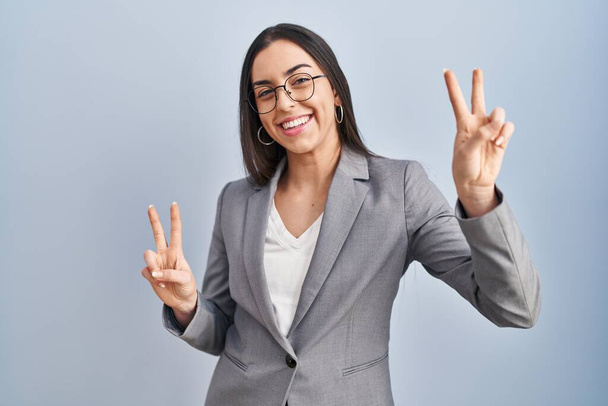 Ισπανίδα επιχειρηματίας που φοράει γυαλιά χαμογελώντας στην κάμερα και δείχνει τα δάχτυλα να κάνουν το σύμβολο της νίκης. Νούμερο δύο..  - Φωτογραφία, εικόνα