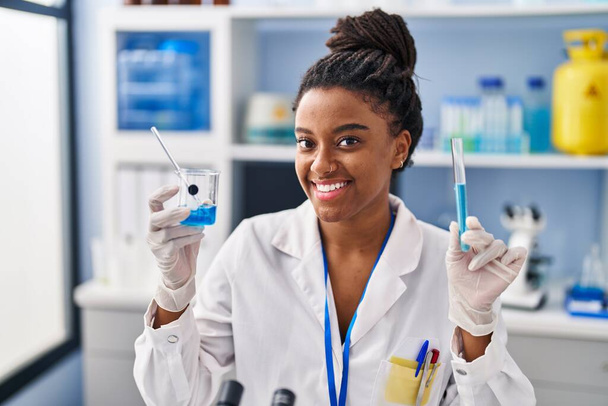 афроамериканська жінка-науковець посміхається впевнено тримає пробірку і скло в лабораторії - Фото, зображення