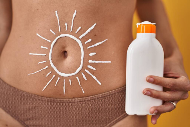 jonge vrouw dragen bikini met zon lotion tekenen op buik over geel geïsoleerde achtergrond - Foto, afbeelding