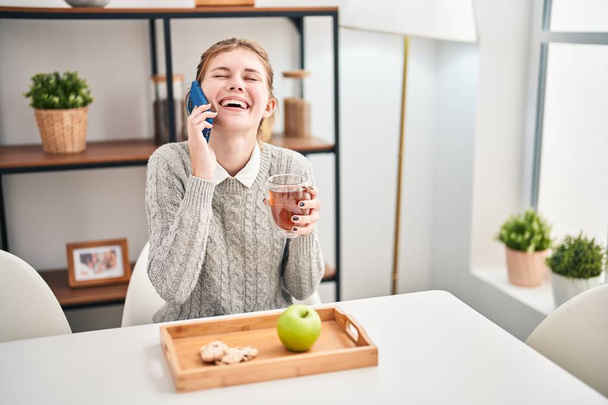 Sağlıklı kahvaltı yapan genç sarışın kadın yemek odasında akıllı telefondan konuşuyor. - Fotoğraf, Görsel