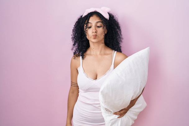 Hispanic woman with curly hair wearing sleep mask and pajama holding pillow making fish face with lips, crazy and comical gesture. funny expression.  - Photo, Image
