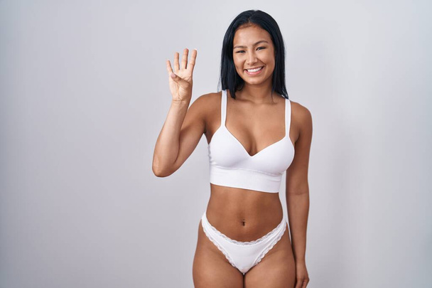 Mujer hispana vistiendo lencería mostrando y señalando con los dedos el número cuatro mientras sonríe confiada y feliz.  - Foto, Imagen