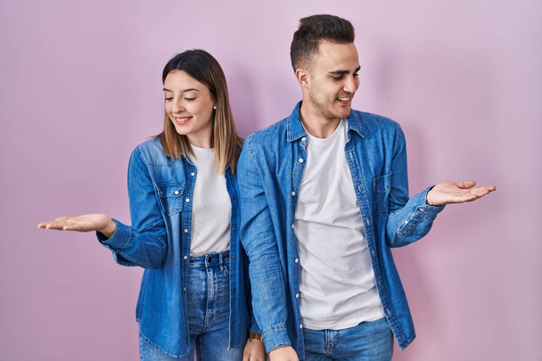ピンクの背景の上に立っている若いヒスパニックカップルは,両手を開いた手のひらを示し,提示し,広告の比較とバランス  - 写真・画像