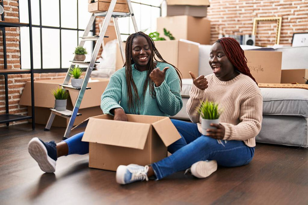 Dos mujeres africanas se mudan a una nueva casa sentadas en el suelo apuntando con el pulgar hacia el costado sonriendo felices con la boca abierta  - Foto, Imagen