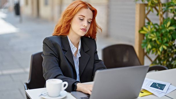 Молода руда жінка бізнесмен використовує ноутбук з розслабленим виразом на терасі кав'ярні
 - Фото, зображення