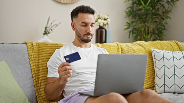 młody arabski człowiek zakupy z laptopem i karty kredytowej siedzi na kanapie w domu - Zdjęcie, obraz