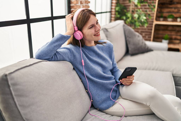 Giovane donna bionda che ascolta musica seduta sul divano di casa - Foto, immagini