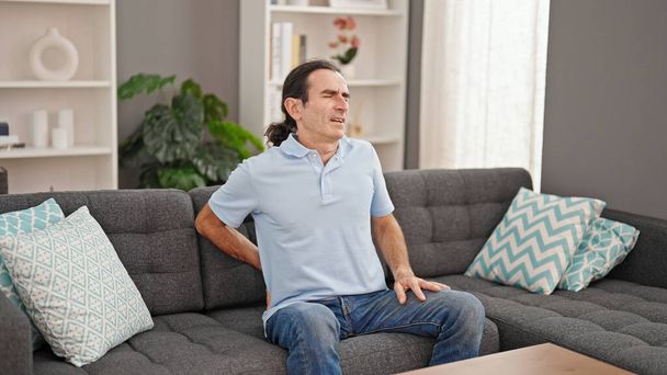 Μεσήλικας άνθρωπος που υποφέρει για πόνο στην πλάτη κάθεται στον καναπέ στο σπίτι - Φωτογραφία, εικόνα