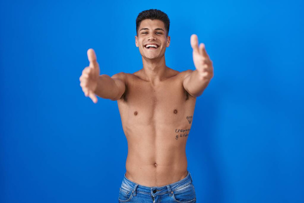 Junger hispanischer Mann steht hemdlos vor blauem Hintergrund und blickt lächelnd mit offenen Armen in die Kamera, um sich zu umarmen. fröhlicher Ausdruck, der das Glück umarmt.  - Foto, Bild