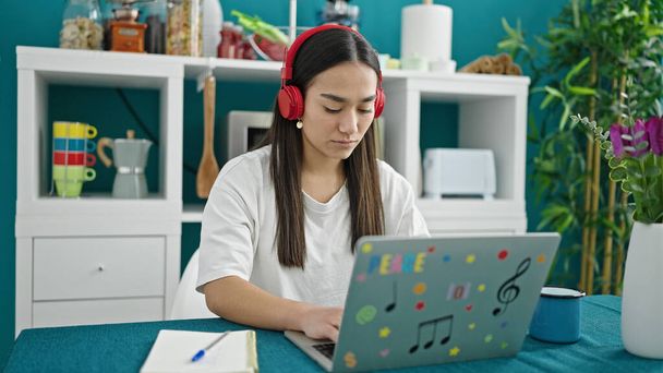 Young beautiful hispanic woman using laptop and headphones sitting on table at dinning room - Photo, Image