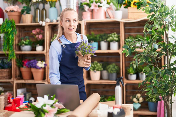 Young blonde woman florist using laptop holding lavender plant at florist - 写真・画像