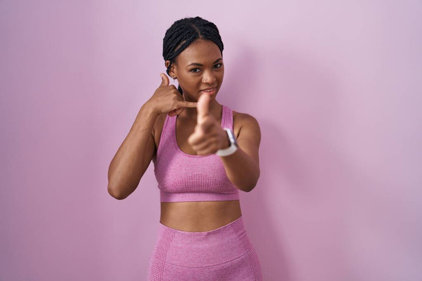 Afro-Amerikaanse vrouw met vlechten dragen sportkleding over roze achtergrond glimlachend doen praten op de telefoon gebaar en wijzen naar u. Bel me maar..  - Foto, afbeelding