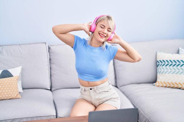 Jeune femme blonde dansant et écoutant de la musique assise sur le canapé à la maison - Photo, image