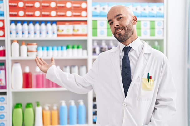 Fiatal kopasz férfi gyógyszerész mosolygós magabiztos áll a gyógyszertárban - Fotó, kép