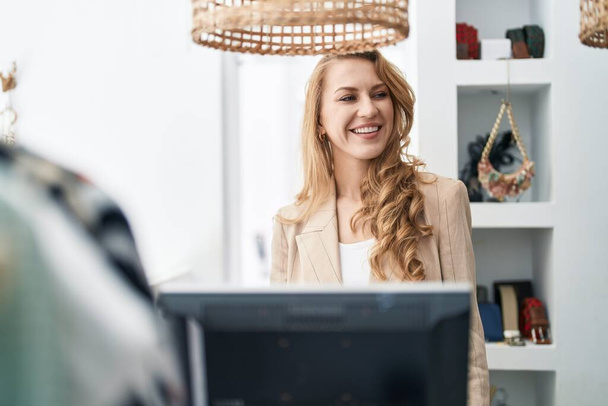 Mujer rubia joven asistente de tienda sonriendo confiado trabajando en la tienda de ropa - Foto, imagen